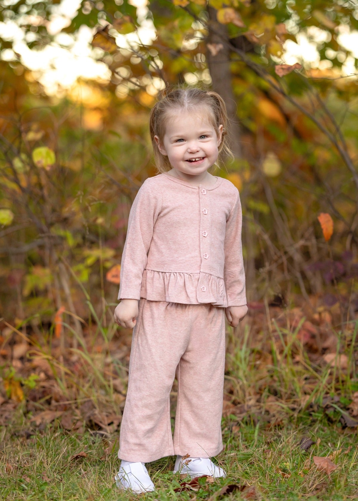 A young child stands and smiles outdoors among green and brown foliage, wearing the Mabel and Honey Margret Long Sleeve Solid Peplum Ruffle Hem Top and Pant Set in heathered pink. The charming ensemble features a peplum ruffle hem top paired with matching pants, adding a sweet touch to the look. Grass and trees provide a natural background.