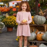 A young girl with curly red hair stands smiling in a pumpkin patch, wearing the Mabel and Honey Margret Long Sleeve Solid Button Front Dropwaist Dress in heathered pink. The dress features flower-shaped buttons and a charming dropwaist silhouette. She holds a small striped gourd, surrounded by pumpkins, squash, and yellow flowers.