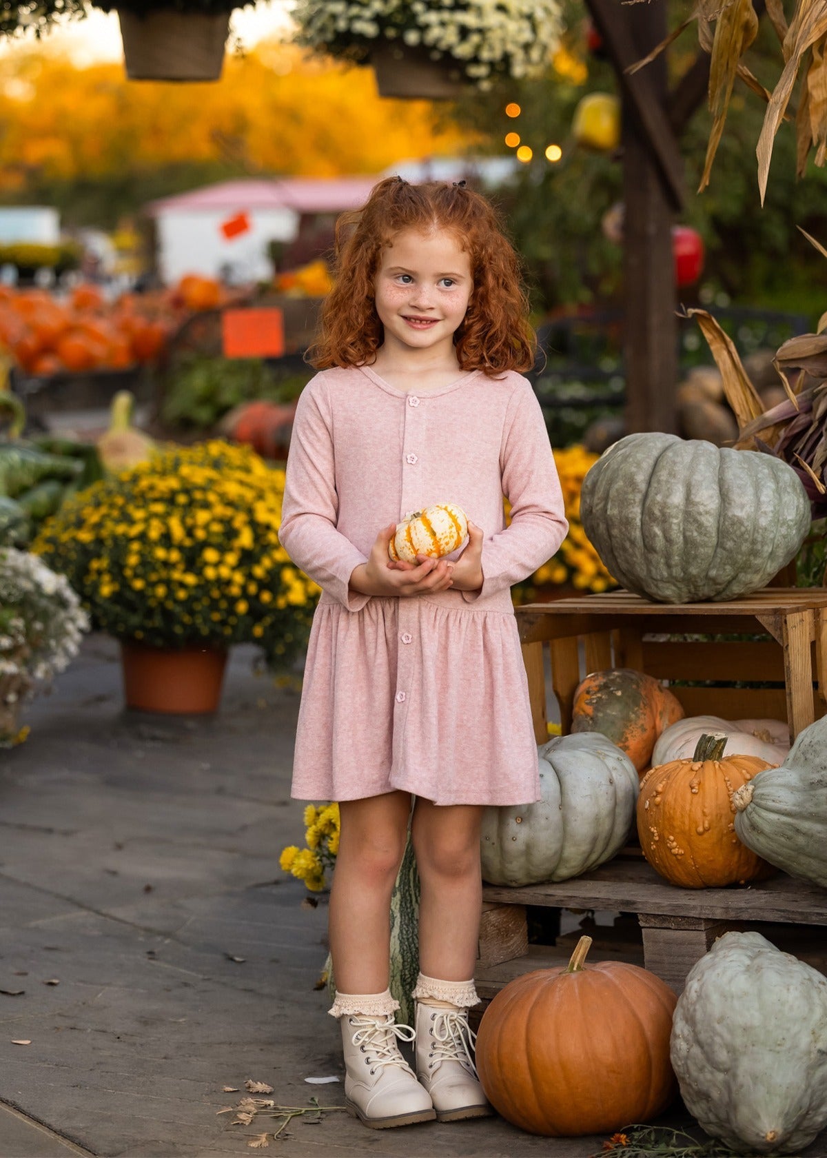A young girl with curly red hair stands smiling in a pumpkin patch, wearing the Mabel and Honey Margret Long Sleeve Solid Button Front Dropwaist Dress in heathered pink. The dress features flower-shaped buttons and a charming dropwaist silhouette. She holds a small striped gourd, surrounded by pumpkins, squash, and yellow flowers.