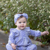 A baby sits barefoot on a path surrounded by white flowering bushes, smiling and reaching out with one hand, while wearing the Mabel and Honey Lavender Lane Long Sleeve Print Ruffle Neck Bubble Romper with a matching headband—adding an extra touch of sweetness to her joyful moment.