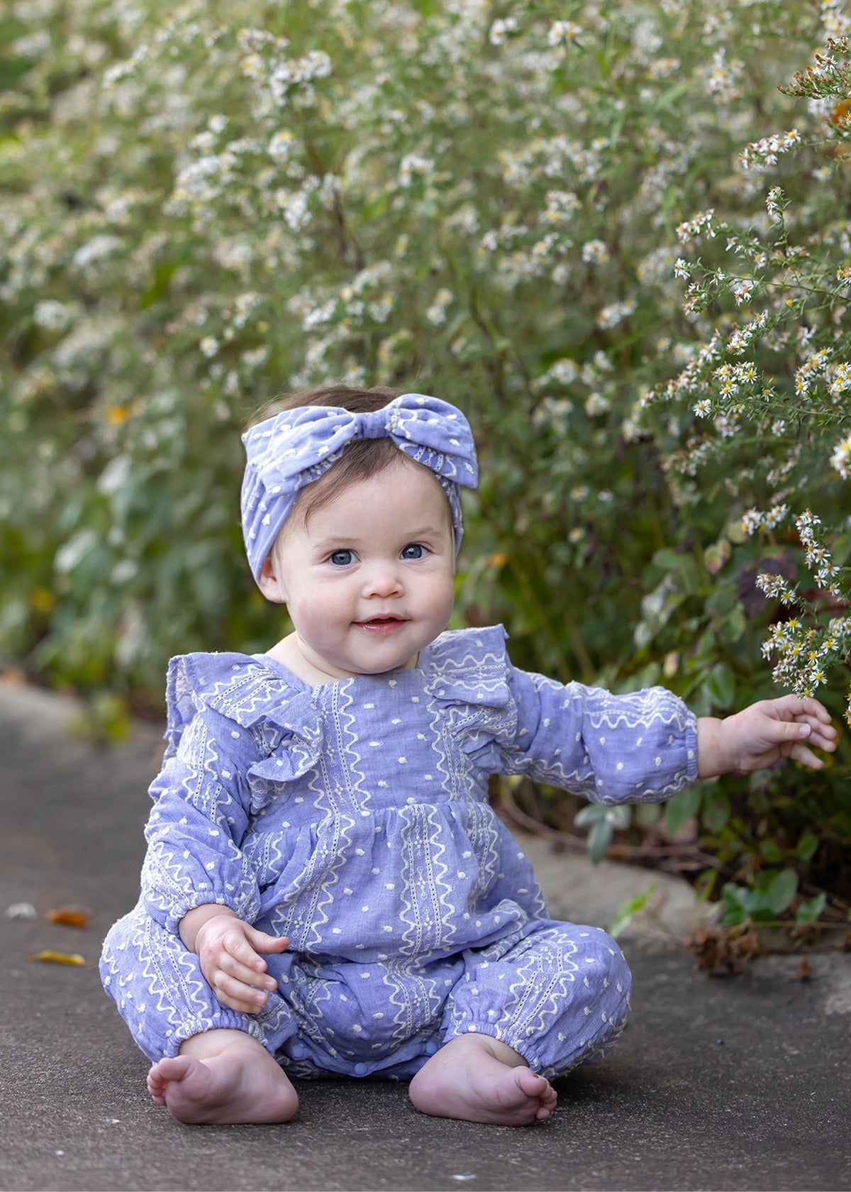 A baby sits barefoot on a path surrounded by white flowering bushes, smiling and reaching out with one hand, while wearing the Mabel and Honey Lavender Lane Long Sleeve Print Ruffle Neck Bubble Romper with a matching headband—adding an extra touch of sweetness to her joyful moment.