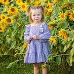 A young girl, her hair in two pigtails, stands smiling among tall sunflowers in a field wearing the Lavender Lane Long Sleeve Embroidered Ruffle Trim Tiered Dress by Mabel and Honey.