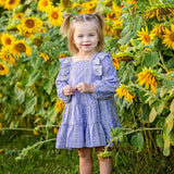 A young girl, her hair in two pigtails, stands smiling among tall sunflowers in a field wearing the Lavender Lane Long Sleeve Embroidered Ruffle Trim Tiered Dress by Mabel and Honey.