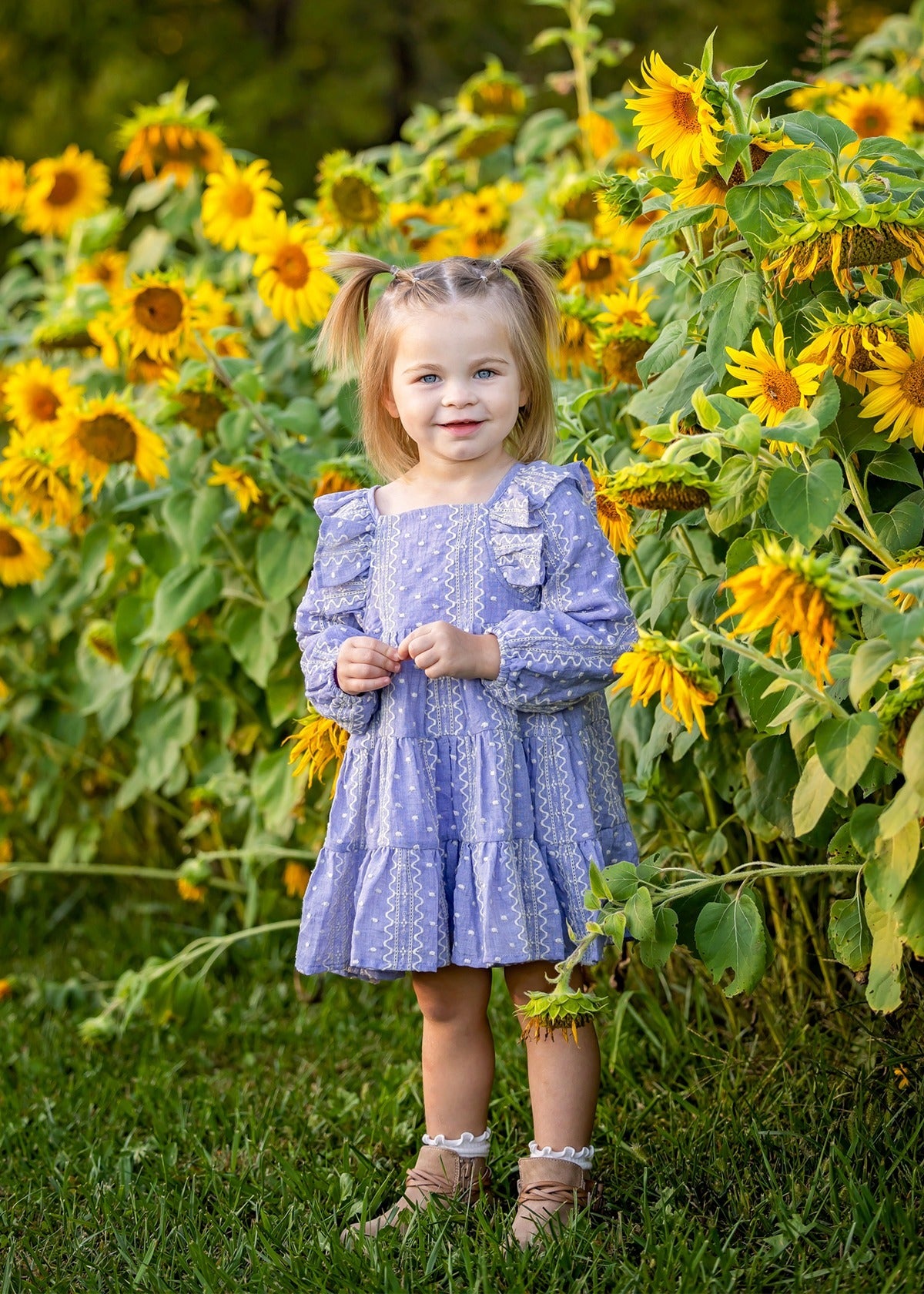 A young girl, her hair in two pigtails, stands smiling among tall sunflowers in a field wearing the Lavender Lane Long Sleeve Embroidered Ruffle Trim Tiered Dress by Mabel and Honey.