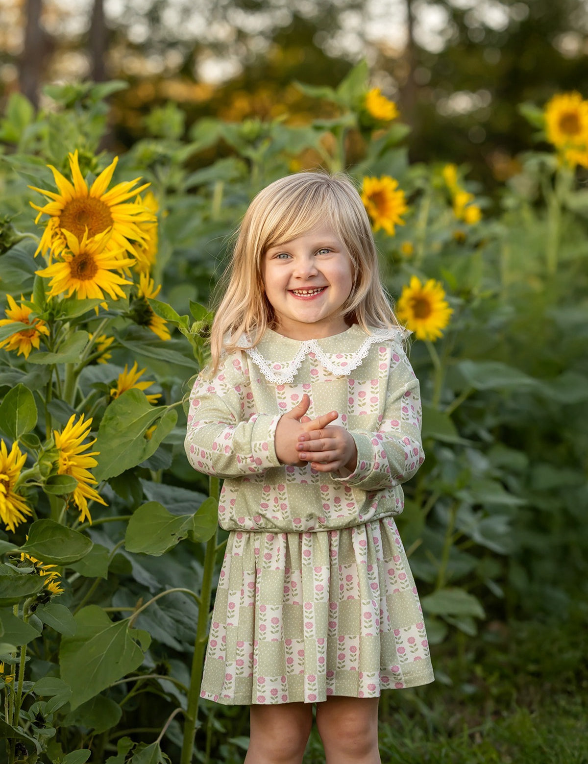 A young girl wearing the Mabel and Honey Olive Long Sleeve Floral Peter Pan Collar Top and Skirt Set smiles as she stands among tall sunflowers in a green field, with trees softly blurred in the background.