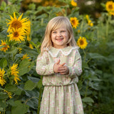 A young girl wearing the Mabel and Honey Olive Long Sleeve Floral Peter Pan Collar Top and Skirt Set smiles as she stands among tall sunflowers in a green field, with trees softly blurred in the background.