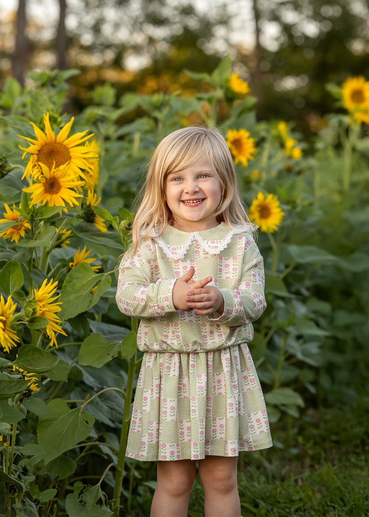 A young girl wearing the Mabel and Honey Olive Long Sleeve Floral Peter Pan Collar Top and Skirt Set smiles as she stands among tall sunflowers in a green field, with trees softly blurred in the background.