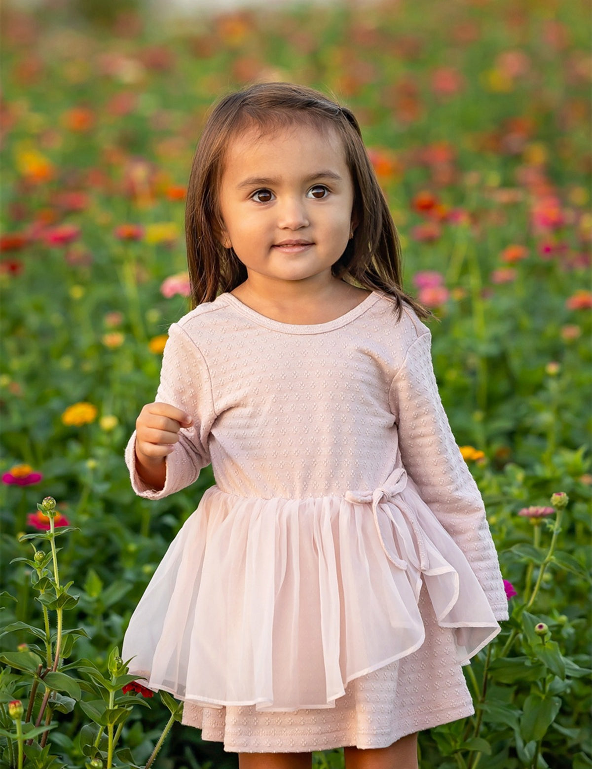 A young girl wearing the Emma Long Sleeve Textured Tiered Tulle Skirt Dress by Mabel and Honey stands outdoors in a field of colorful flowers, looking slightly to the side with a gentle expression.