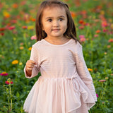 A young girl wearing the Emma Long Sleeve Textured Tiered Tulle Skirt Dress by Mabel and Honey stands outdoors in a field of colorful flowers, looking slightly to the side with a gentle expression.