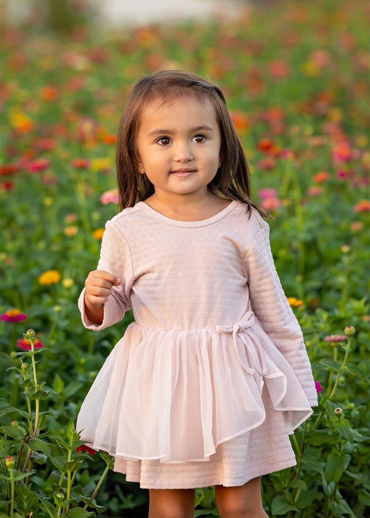 A young girl wearing the Emma Long Sleeve Textured Tiered Tulle Skirt Dress by Mabel and Honey stands outdoors in a field of colorful flowers, looking slightly to the side with a gentle expression.
