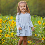 A young girl stands smiling in a sunflower field with green trees and warm sunlight in the background, wearing the Mabel and Honey Eva Long Sleeve Textured Dot Bow Trim Fit and Flare Dress paired with brown boots.