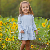 A young girl stands smiling in a sunflower field with green trees and warm sunlight in the background, wearing the Mabel and Honey Eva Long Sleeve Textured Dot Bow Trim Fit and Flare Dress paired with brown boots.