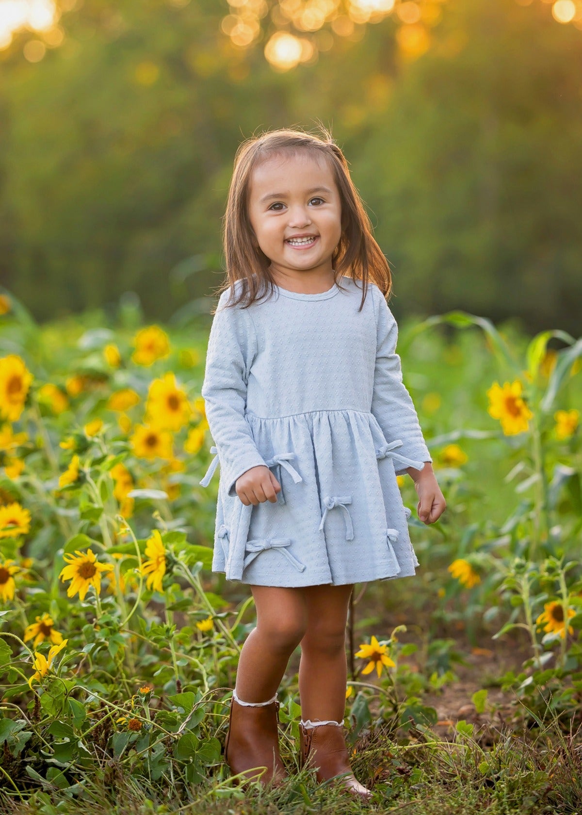 A young girl stands smiling in a sunflower field with green trees and warm sunlight in the background, wearing the Mabel and Honey Eva Long Sleeve Textured Dot Bow Trim Fit and Flare Dress paired with brown boots.