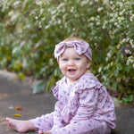 Wearing the Mabel and Honey Victoria Long Sleeve Floral Ruffle Shoulder Straight Leg Romper and a coordinating headband, a smiling baby sits on a sidewalk—soft mauve florals setting the tone for a perfect baby girl fall outfit—surrounded by green foliage and delicate white flowers in the background.