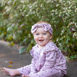 Wearing the Mabel and Honey Victoria Long Sleeve Floral Ruffle Shoulder Straight Leg Romper and a coordinating headband, a smiling baby sits on a sidewalk—soft mauve florals setting the tone for a perfect baby girl fall outfit—surrounded by green foliage and delicate white flowers in the background.