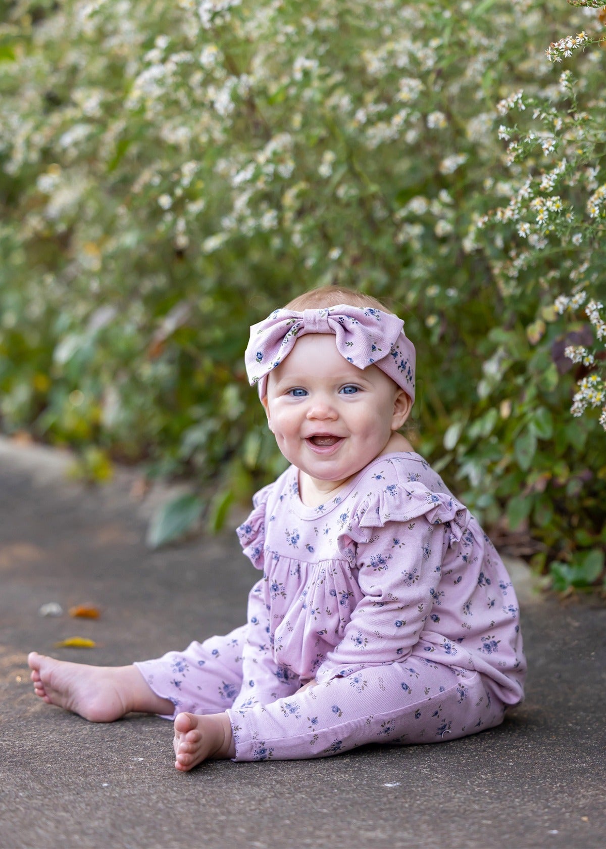 Wearing the Mabel and Honey Victoria Long Sleeve Floral Ruffle Shoulder Straight Leg Romper and a coordinating headband, a smiling baby sits on a sidewalk—soft mauve florals setting the tone for a perfect baby girl fall outfit—surrounded by green foliage and delicate white flowers in the background.