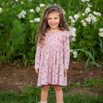 A young girl with long curly hair smiles while standing in front of a green field with blooming white flowers, wearing the Victoria Long Sleeve Floral Ruffle Shoulder Tiered Dress by Mabel and Honey and cream boots.