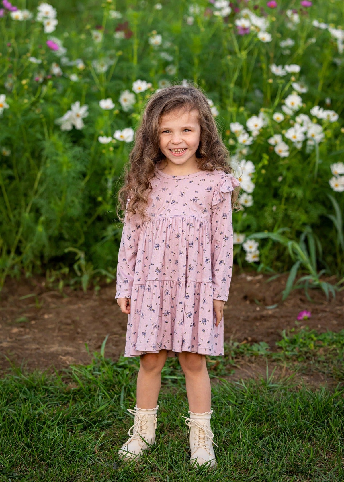 A young girl with long curly hair smiles while standing in front of a green field with blooming white flowers, wearing the Victoria Long Sleeve Floral Ruffle Shoulder Tiered Dress by Mabel and Honey and cream boots.