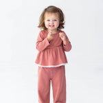 A young child with brown hair smiles, standing in the Mabel and Honey Maribelle Long Sleeve Polka Dot Peplum Top and Pant Set—a girls outfit featuring a peplum top and wide-leg pants in dusty rose—paired with white shoes against a plain white background.