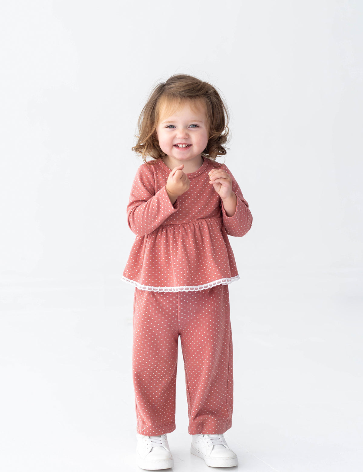 A young child with brown hair smiles, standing in the Mabel and Honey Maribelle Long Sleeve Polka Dot Peplum Top and Pant Set—a girls outfit featuring a peplum top and wide-leg pants in dusty rose—paired with white shoes against a plain white background.