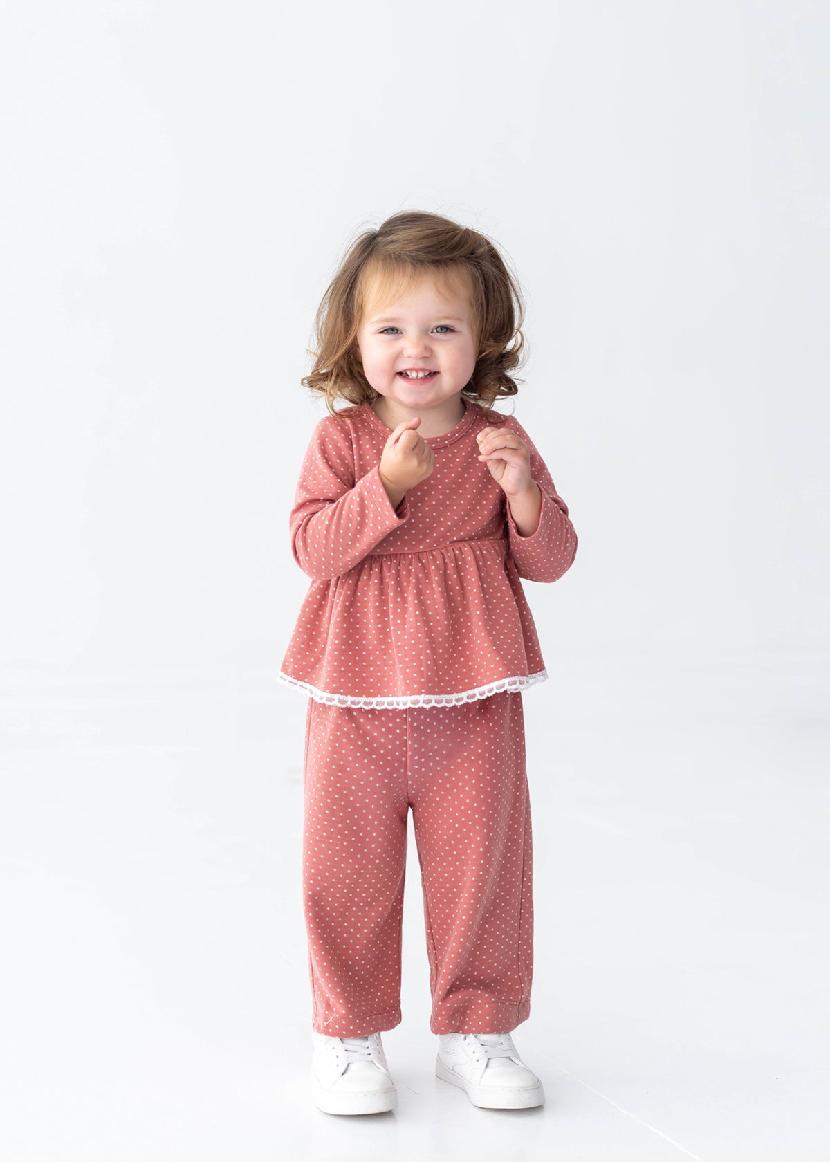 A young child with brown hair smiles, standing in the Mabel and Honey Maribelle Long Sleeve Polka Dot Peplum Top and Pant Set—a girls outfit featuring a peplum top and wide-leg pants in dusty rose—paired with white shoes against a plain white background.