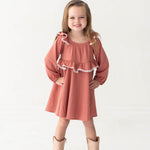 A young girl with long hair smiles confidently, wearing the Maribelle Bishop Sleeve Polka Dot Ruffle Collar A Line Dress by Mabel and Honey and tan cowboy boots, standing against a plain white background.