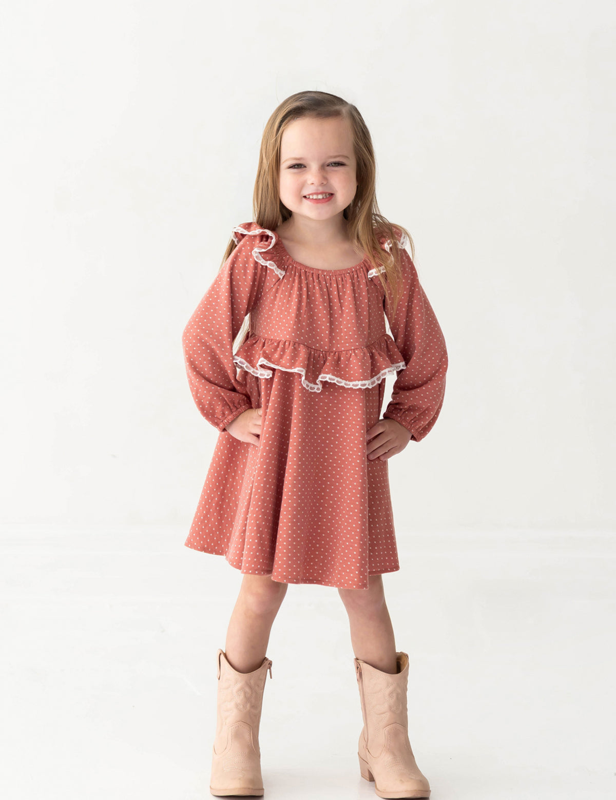 A young girl with long hair smiles confidently, wearing the Maribelle Bishop Sleeve Polka Dot Ruffle Collar A Line Dress by Mabel and Honey and tan cowboy boots, standing against a plain white background.