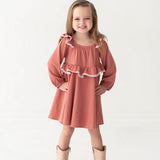 A young girl with long hair smiles confidently, wearing the Maribelle Bishop Sleeve Polka Dot Ruffle Collar A Line Dress by Mabel and Honey and tan cowboy boots, standing against a plain white background.