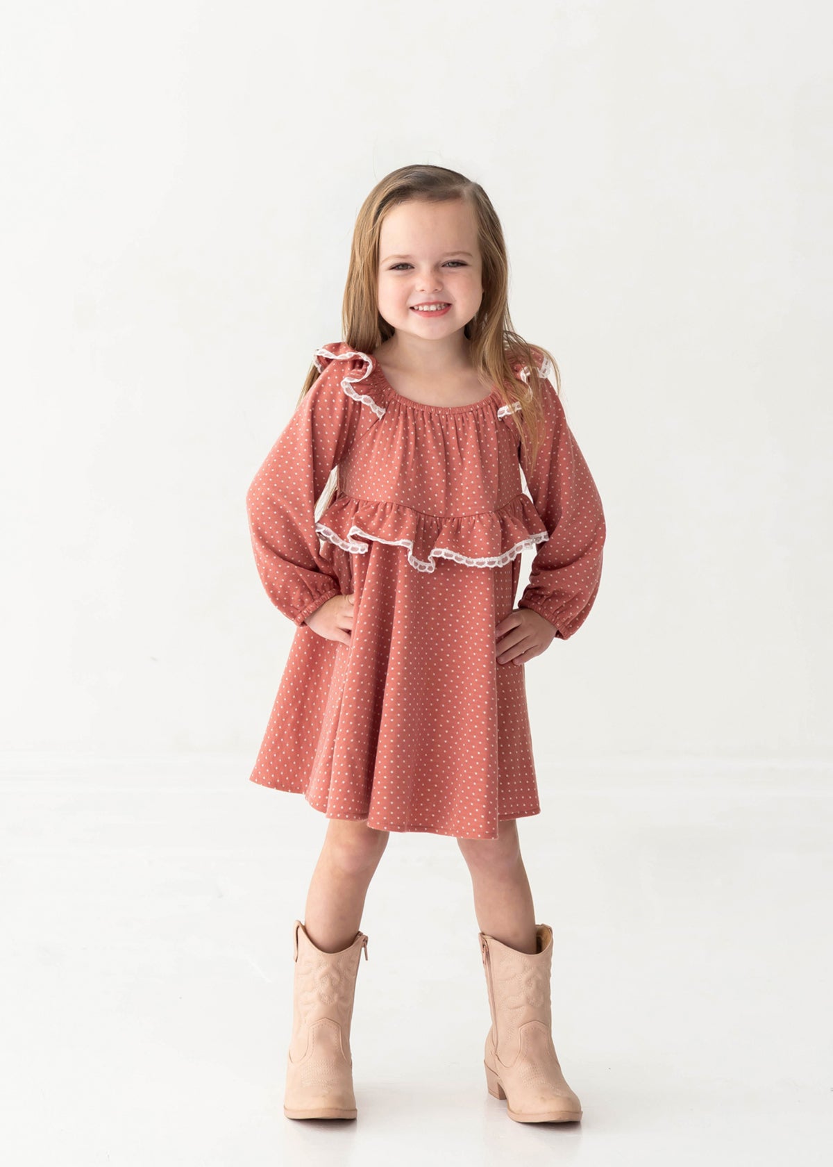 A young girl with long hair smiles confidently, wearing the Maribelle Bishop Sleeve Polka Dot Ruffle Collar A Line Dress by Mabel and Honey and tan cowboy boots, standing against a plain white background.