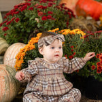 A baby wearing the Mabel and Honey Chestnut Long Sleeve Plaid Ruffle Collar Smocked Bubble Romper with a matching headband sits on straw surrounded by pumpkins and vibrant autumn blooms, including red and yellow mums, in a festive fall scene.