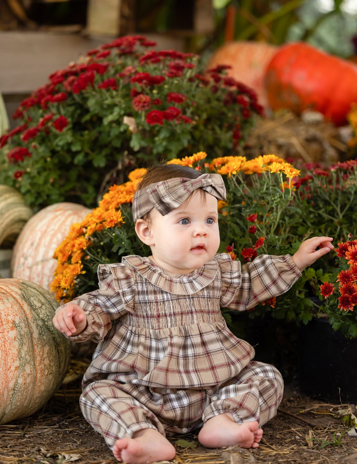 A baby wearing the Mabel and Honey Chestnut Long Sleeve Plaid Ruffle Collar Smocked Bubble Romper with a matching headband sits on straw surrounded by pumpkins and vibrant autumn blooms, including red and yellow mums, in a festive fall scene.