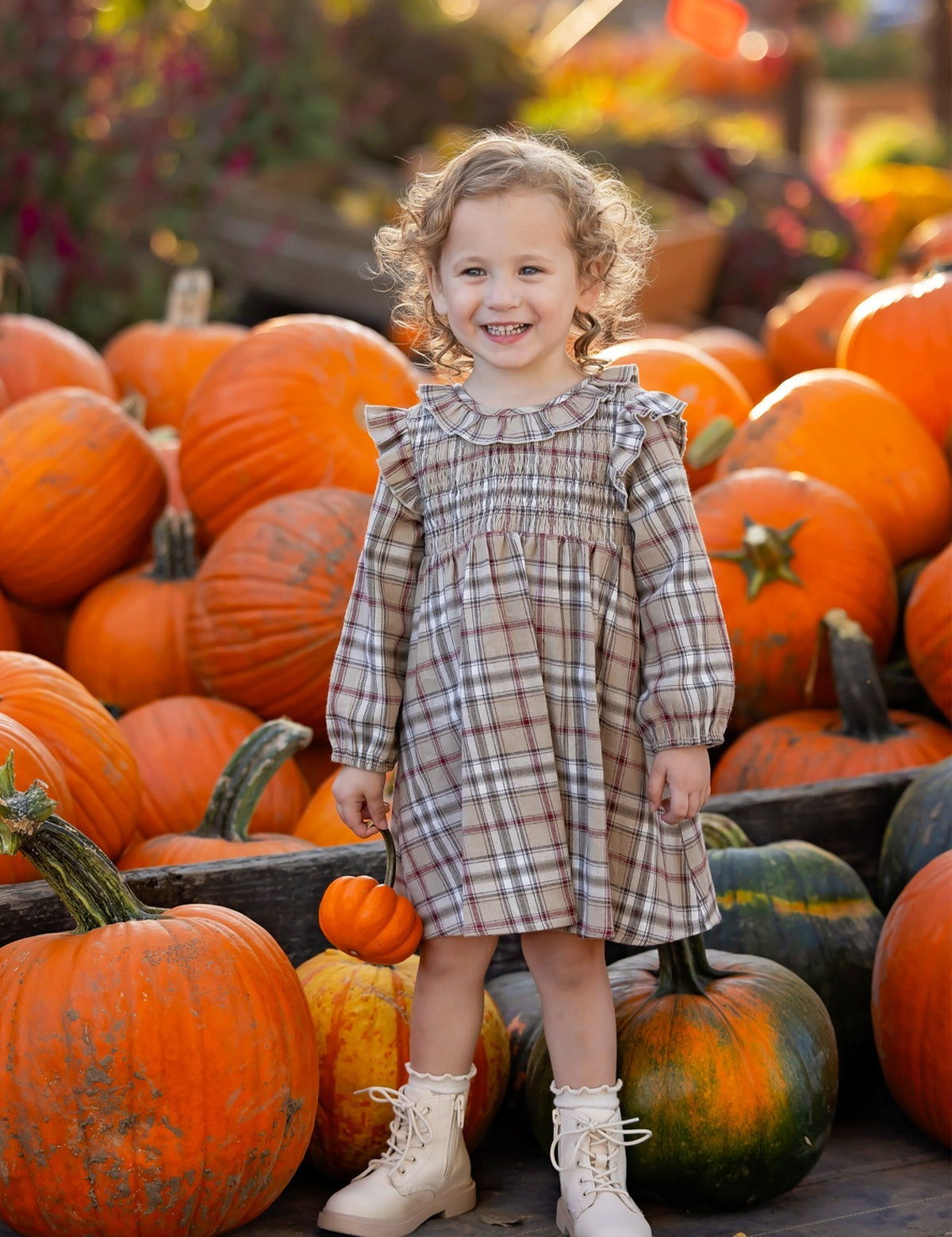A smiling young child wearing the Mabel and Honey Chestnut Long Sleeve Plaid Ruffle Collar Smocked Bodice Dress with boots stands among large orange pumpkins at a pumpkin patch, surrounded by vibrant autumn foliage.