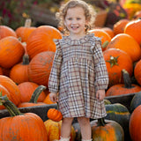 A smiling young child wearing the Mabel and Honey Chestnut Long Sleeve Plaid Ruffle Collar Smocked Bodice Dress with boots stands among large orange pumpkins at a pumpkin patch, surrounded by vibrant autumn foliage.