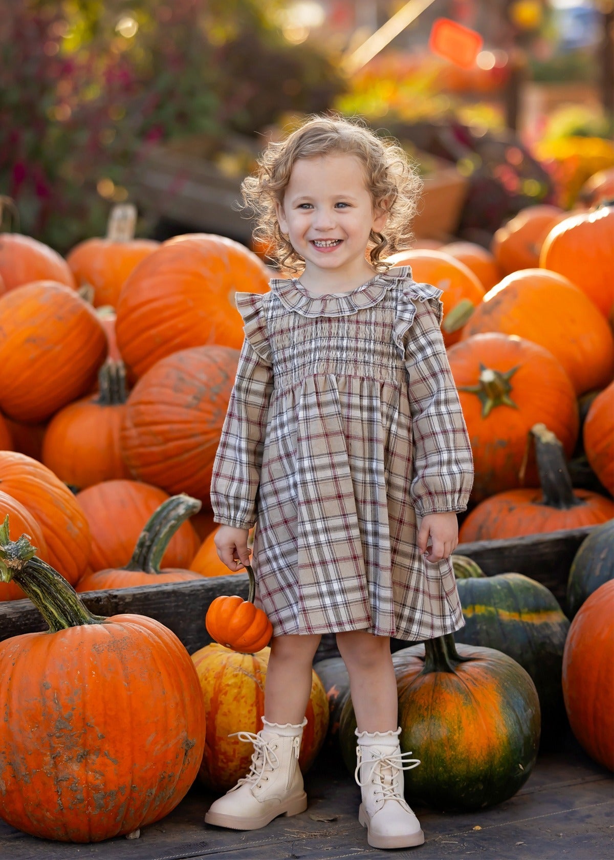 A smiling young child wearing the Mabel and Honey Chestnut Long Sleeve Plaid Ruffle Collar Smocked Bodice Dress with boots stands among large orange pumpkins at a pumpkin patch, surrounded by vibrant autumn foliage.