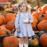 A young girl, smiling at the camera in an outdoor autumn setting, stands among pumpkins wearing the Mabel and Honey Ella Rae Long Sleeve Gingham Peter Pan Collar Tiered Dress and white boots. She holds a small white pumpkin in her hands.