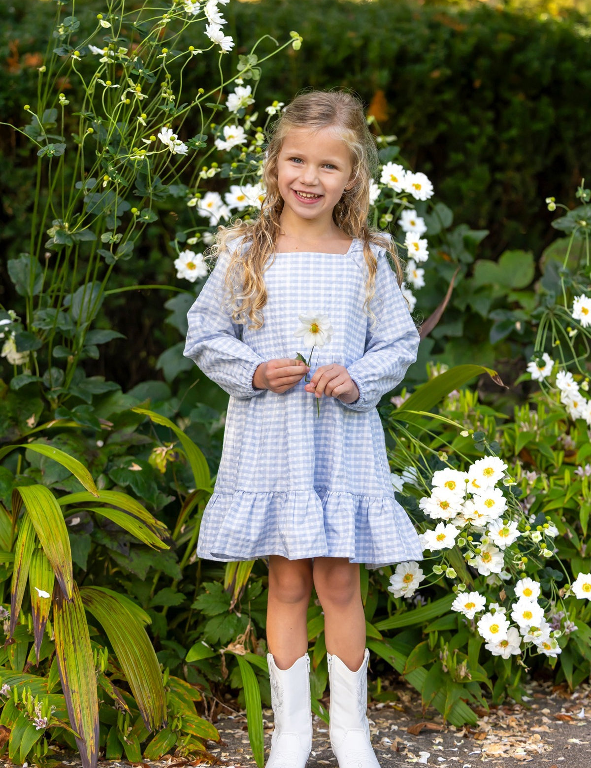 A young girl in the Mabel and Honey Ella Rae Long Sleeve Gingham Ruffle Trim Tiered Dress and white boots stands smiling among tall white flowers, holding a bloom in her hand. Green foliage surrounds her, highlighting the charm of Mabel and Honey’s girls cotton dress collection.