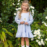 A young girl in the Mabel and Honey Ella Rae Long Sleeve Gingham Ruffle Trim Tiered Dress and white boots stands smiling among tall white flowers, holding a bloom in her hand. Green foliage surrounds her, highlighting the charm of Mabel and Honey’s girls cotton dress collection.