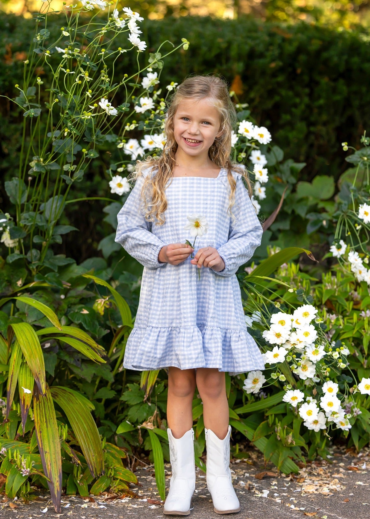 A young girl in the Mabel and Honey Ella Rae Long Sleeve Gingham Ruffle Trim Tiered Dress and white boots stands smiling among tall white flowers, holding a bloom in her hand. Green foliage surrounds her, highlighting the charm of Mabel and Honey’s girls cotton dress collection.