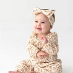 A smiling baby in the Mabel and Honey Autumn Breeze Long Sleeve Floral Ruffle Neck Straight Leg Romper, paired with a matching headband, sits on a white floor with hands raised and gazes slightly to the side—the perfect cozy fall outfit in soft cotton gauze.