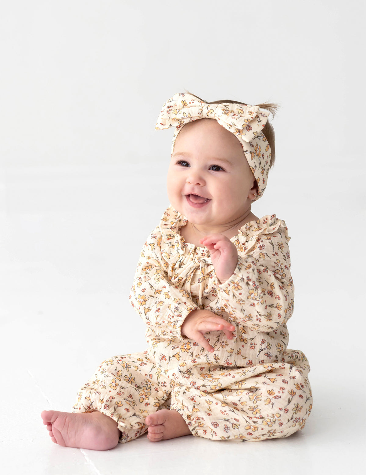 A smiling baby in the Mabel and Honey Autumn Breeze Long Sleeve Floral Ruffle Neck Straight Leg Romper, paired with a matching headband, sits on a white floor with hands raised and gazes slightly to the side—the perfect cozy fall outfit in soft cotton gauze.