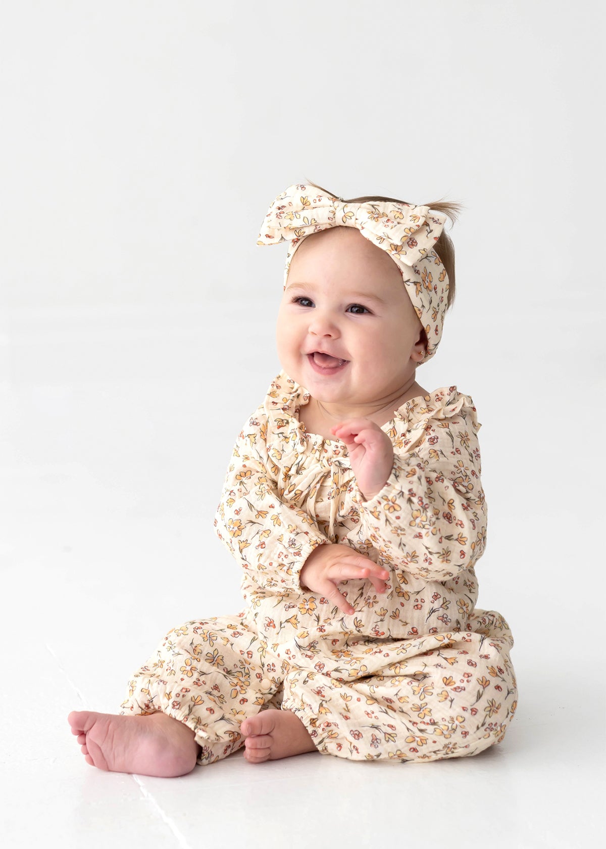 A smiling baby in the Mabel and Honey Autumn Breeze Long Sleeve Floral Ruffle Neck Straight Leg Romper, paired with a matching headband, sits on a white floor with hands raised and gazes slightly to the side—the perfect cozy fall outfit in soft cotton gauze.