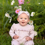 A smiling baby wearing a Mabel and Honey Maggie Long Sleeve Ruffle Trim Straight Leg Romper in heathered lavender, paired with a matching headband, sits on grass in front of blooming pink and white flowers.