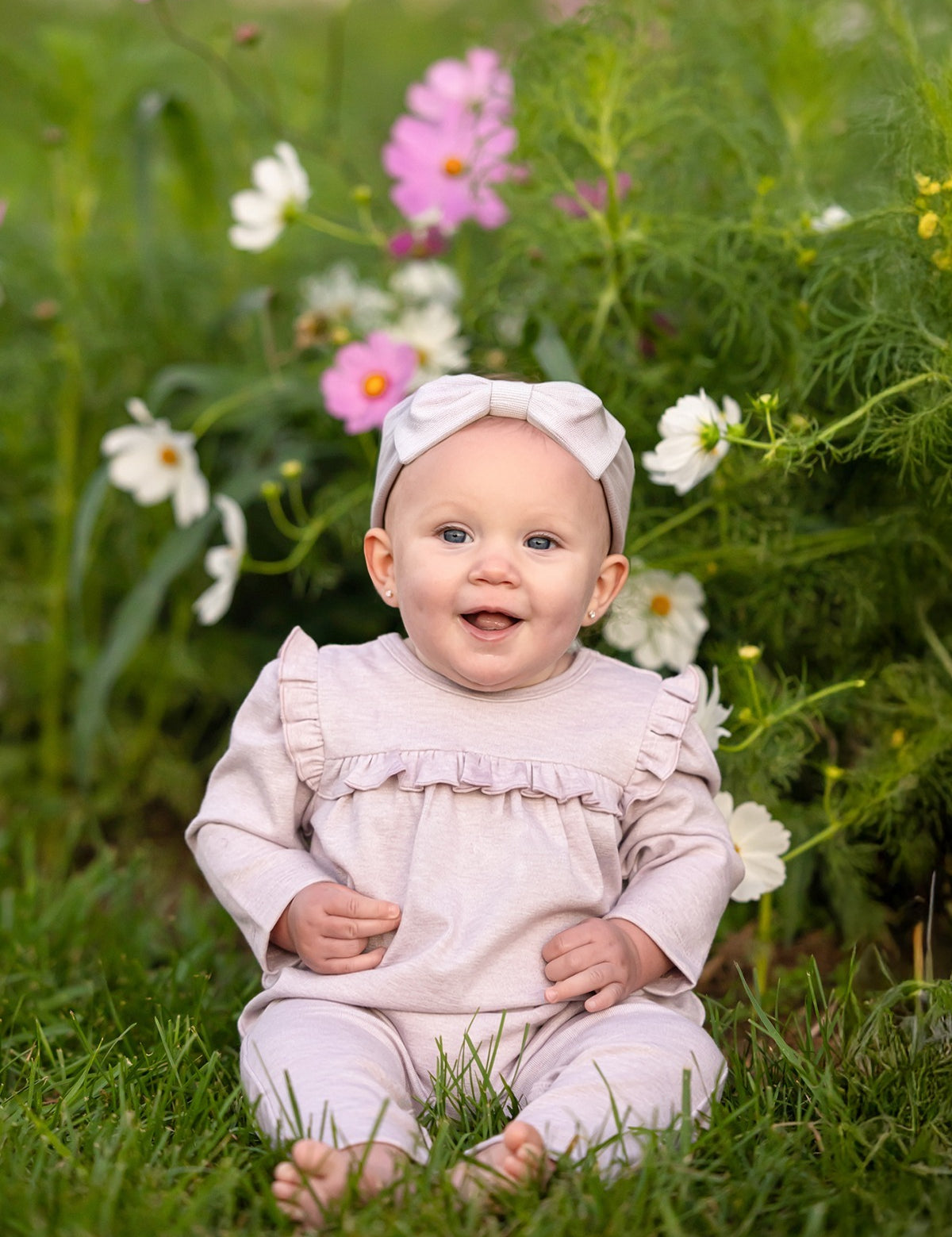 A smiling baby wearing a Mabel and Honey Maggie Long Sleeve Ruffle Trim Straight Leg Romper in heathered lavender, paired with a matching headband, sits on grass in front of blooming pink and white flowers.