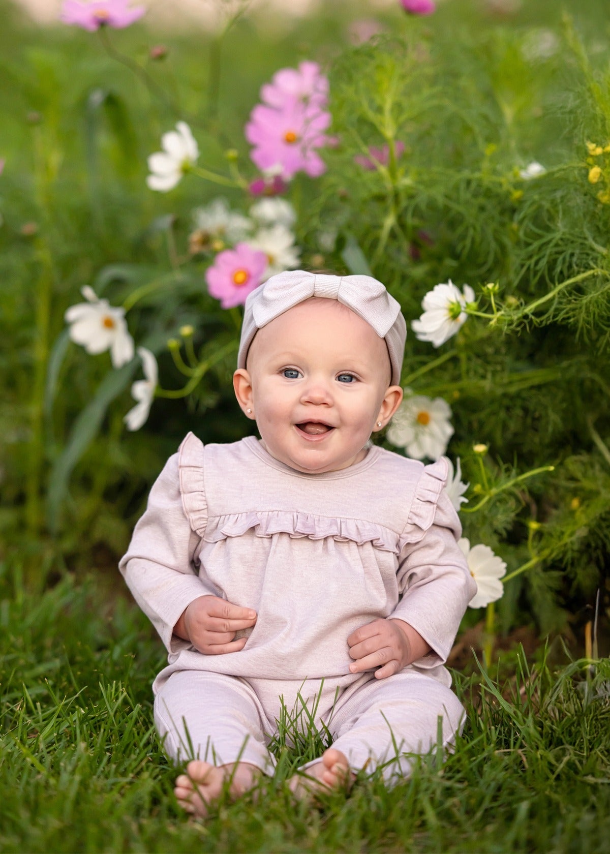 A smiling baby wearing a Mabel and Honey Maggie Long Sleeve Ruffle Trim Straight Leg Romper in heathered lavender, paired with a matching headband, sits on grass in front of blooming pink and white flowers.