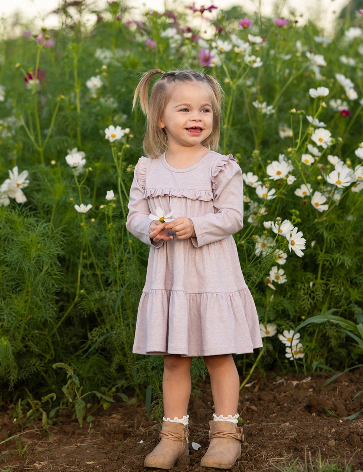 A young girl stands smiling in a field of white and pink flowers, holding a small blossom, wearing the Mabel and Honey Maggie Long Sleeve Ruffle Yoke Tiered Dress in heathered lavender, featuring delicate ruffle trim, with lush green foliage in the background.