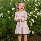 A young girl stands smiling in a field of white and pink flowers, holding a small blossom, wearing the Mabel and Honey Maggie Long Sleeve Ruffle Yoke Tiered Dress in heathered lavender, featuring delicate ruffle trim, with lush green foliage in the background.