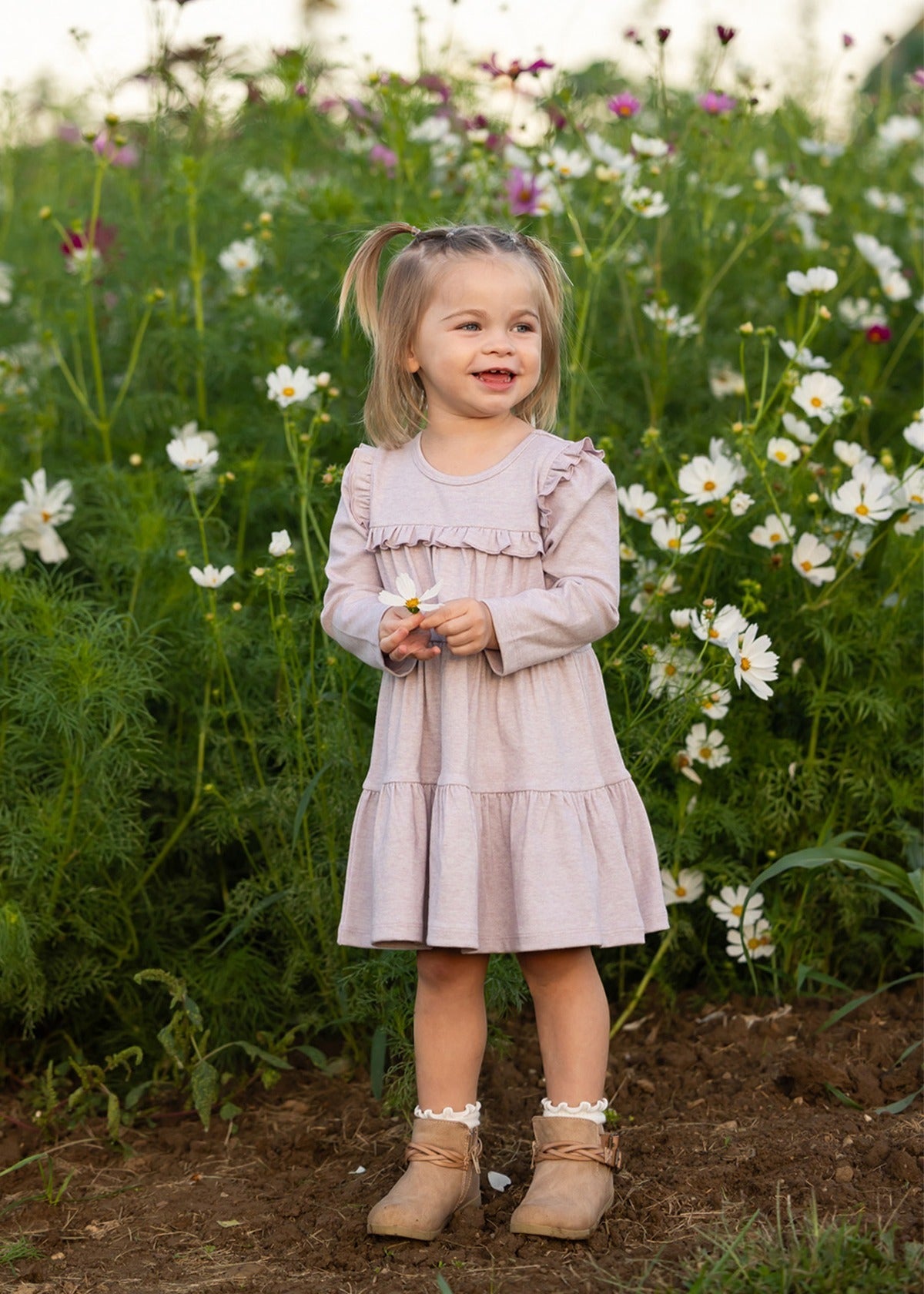 A young girl stands smiling in a field of white and pink flowers, holding a small blossom, wearing the Mabel and Honey Maggie Long Sleeve Ruffle Yoke Tiered Dress in heathered lavender, featuring delicate ruffle trim, with lush green foliage in the background.