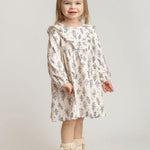 A young girl with light brown hair, wearing the Mabel and Honey Gracie Long Sleeve Floral Ruffle Yoke A Line Dress, cream boots, and a hair clip stands smiling against a plain light background.