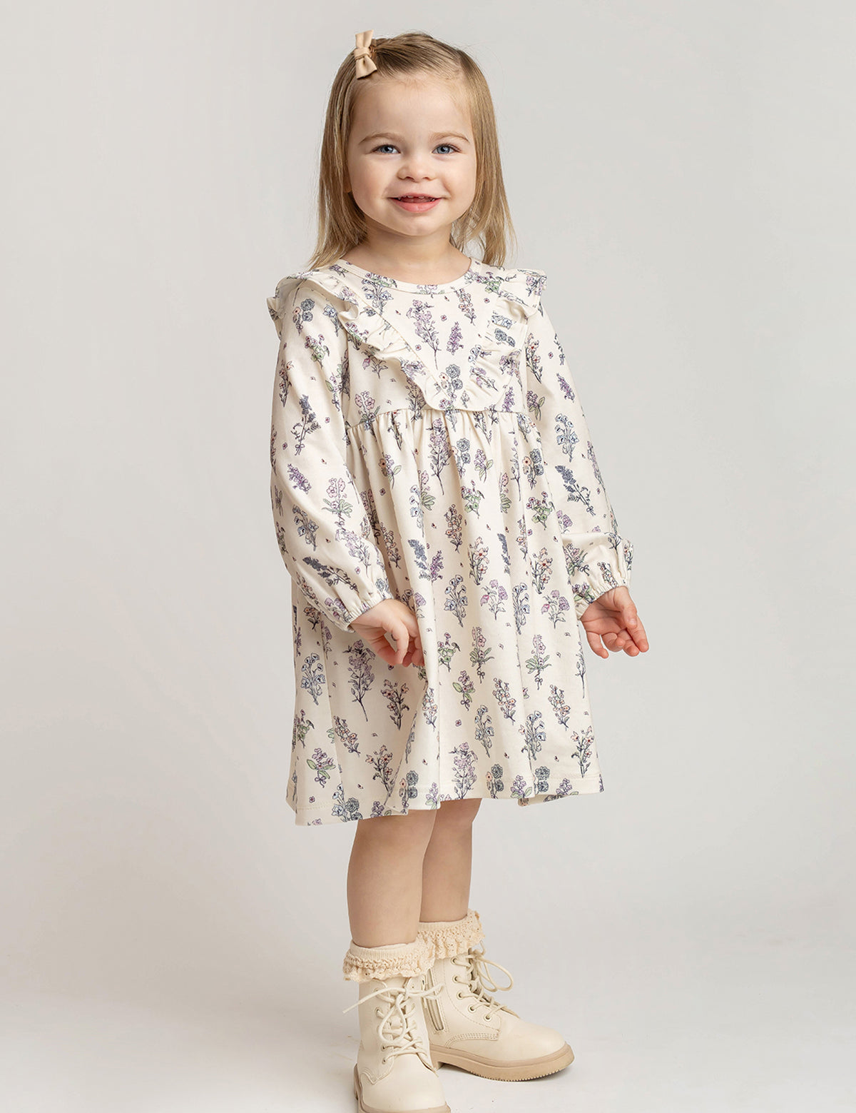 A young girl with light brown hair, wearing the Mabel and Honey Gracie Long Sleeve Floral Ruffle Yoke A Line Dress, cream boots, and a hair clip stands smiling against a plain light background.