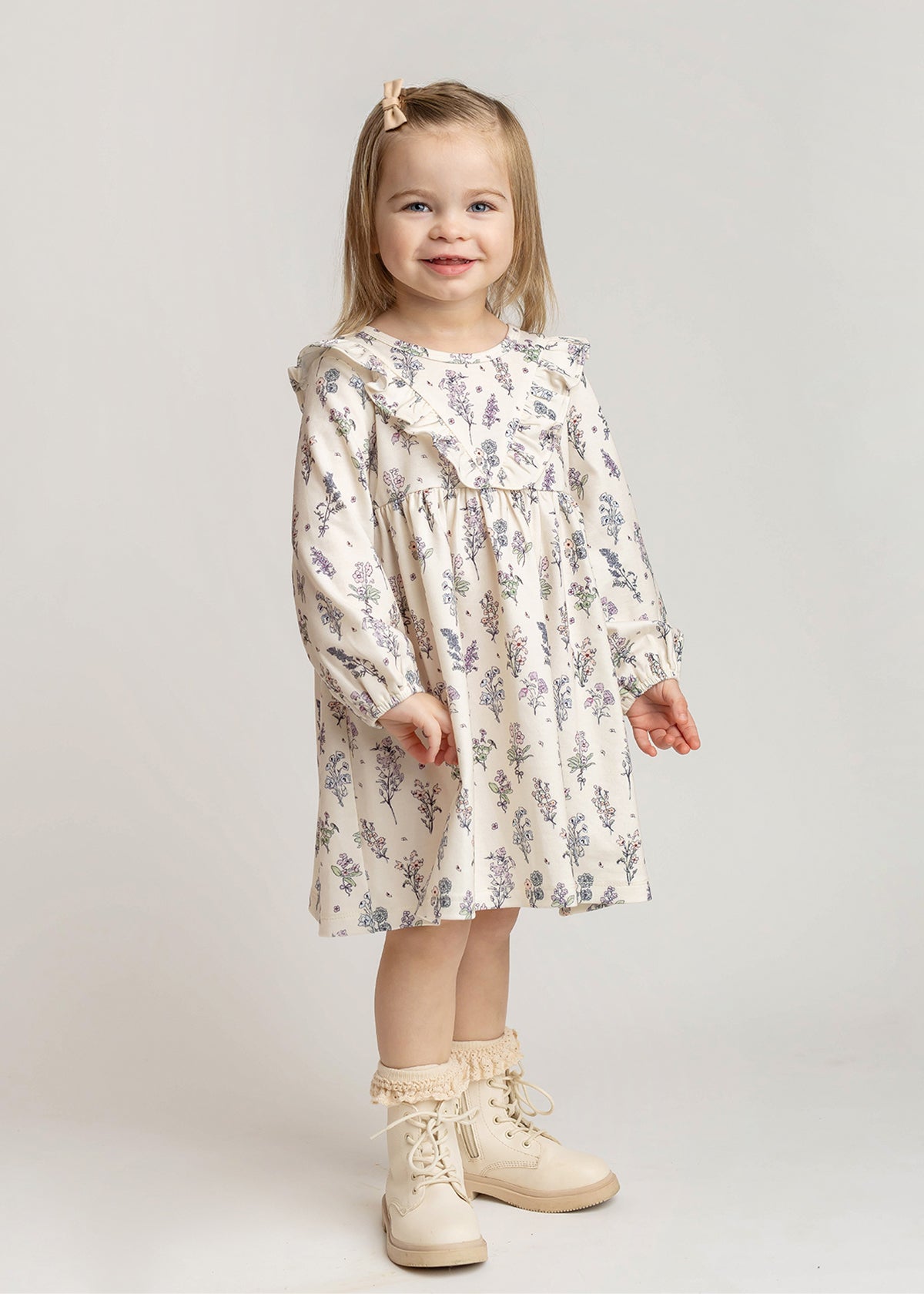 A young girl with light brown hair, wearing the Mabel and Honey Gracie Long Sleeve Floral Ruffle Yoke A Line Dress, cream boots, and a hair clip stands smiling against a plain light background.